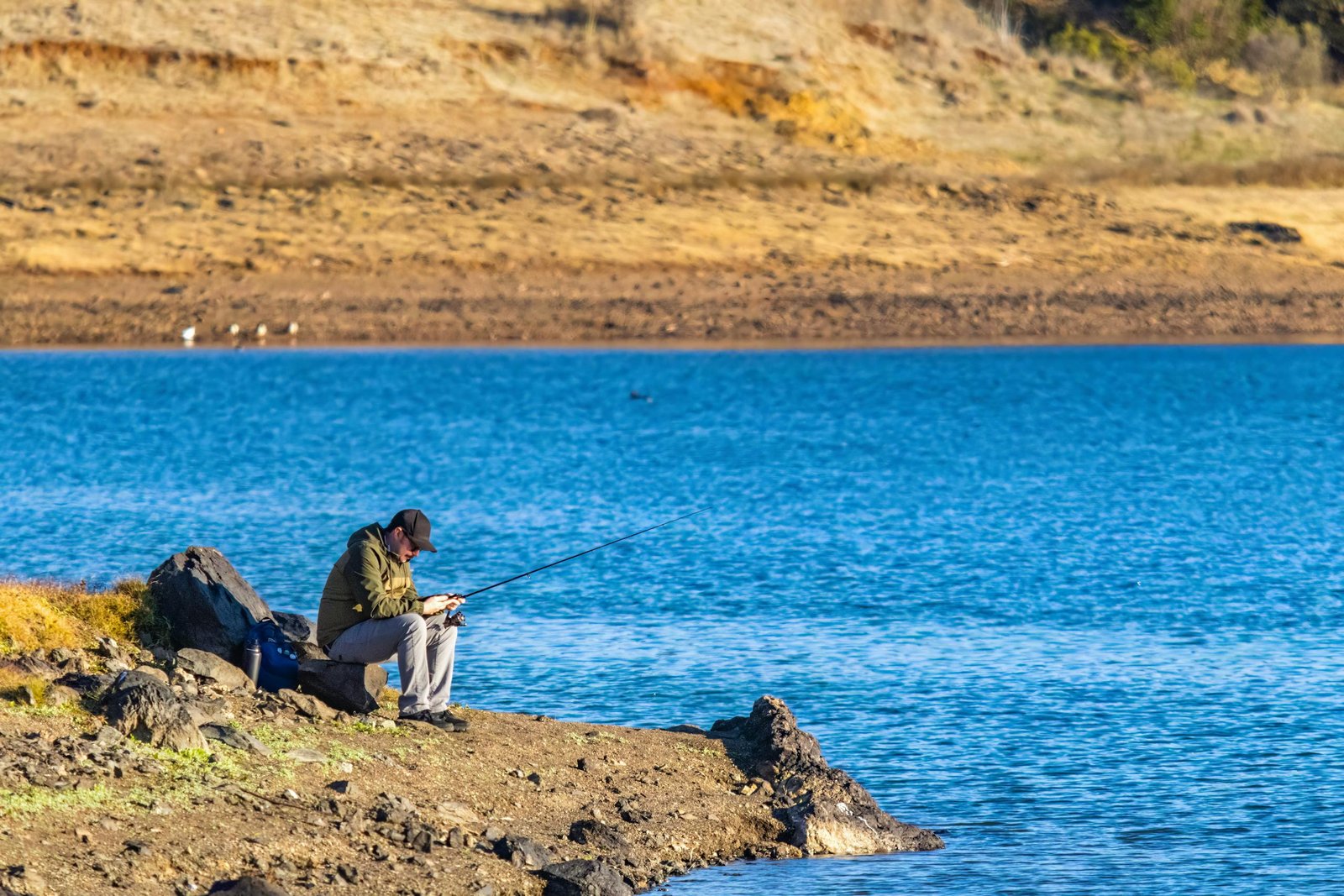 A lone man fishing by the lake on a sunny autumn day, enjoying nature and tranquility.